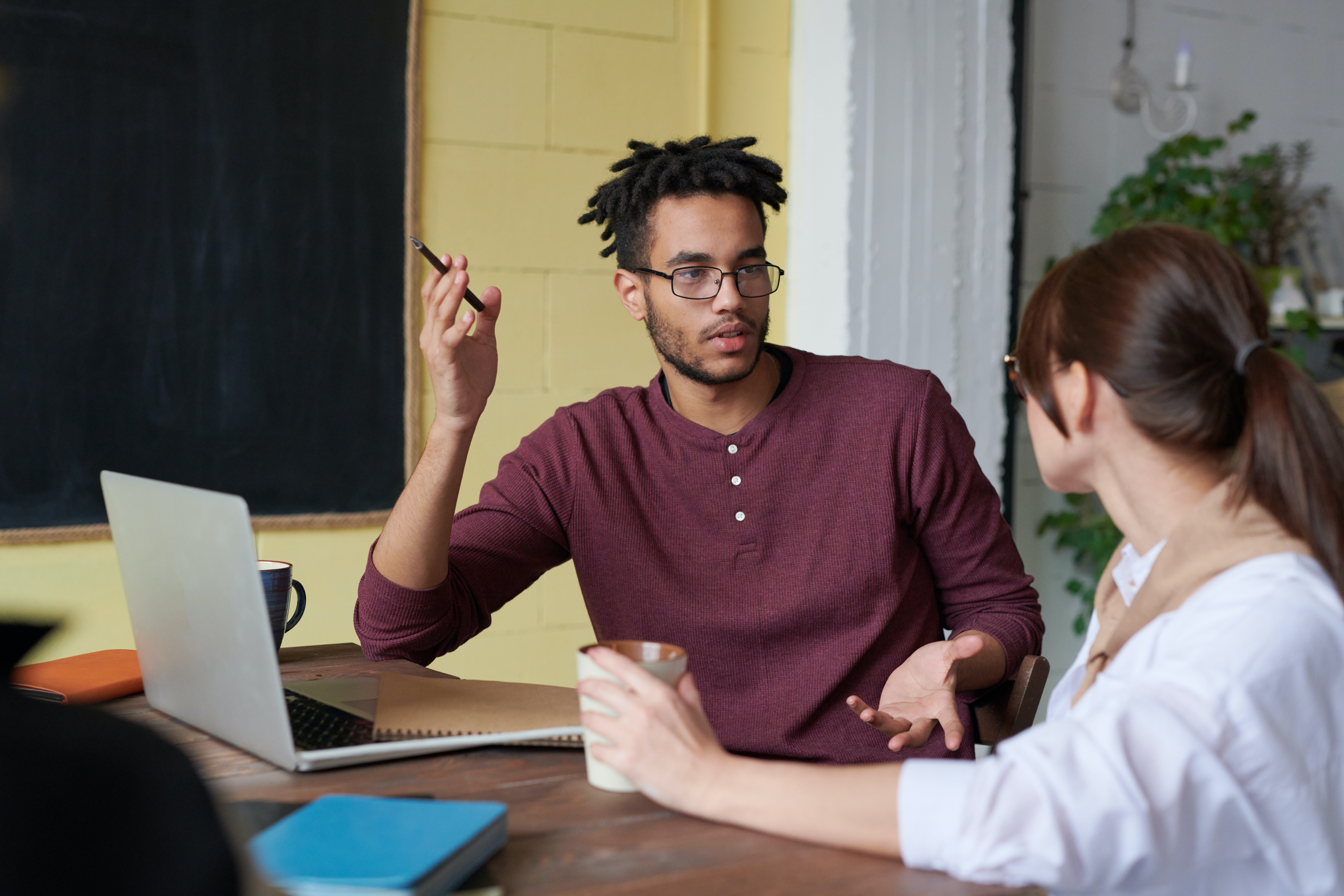 A mixed-race man seated in front of a laptop gesticulates while talking to a young Asian woman.