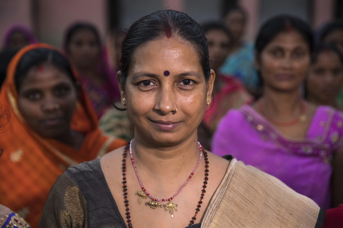 Una mujer líder en Bihar, India se para frente a un grupo, mirando a la cámara con calma confianza y una pequeña sonrisa irónica.
