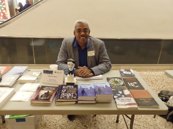 A middle-aged dark-skinned man with glasses and white hair sits at a table displaying his books.