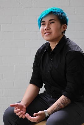 एक एशियाई नॉनबाइनरी ट्रांसमेक्युलिन व्यक्ति अपनी गोद में हाथ रखकर मुस्कुराते हुए बैठता है।
