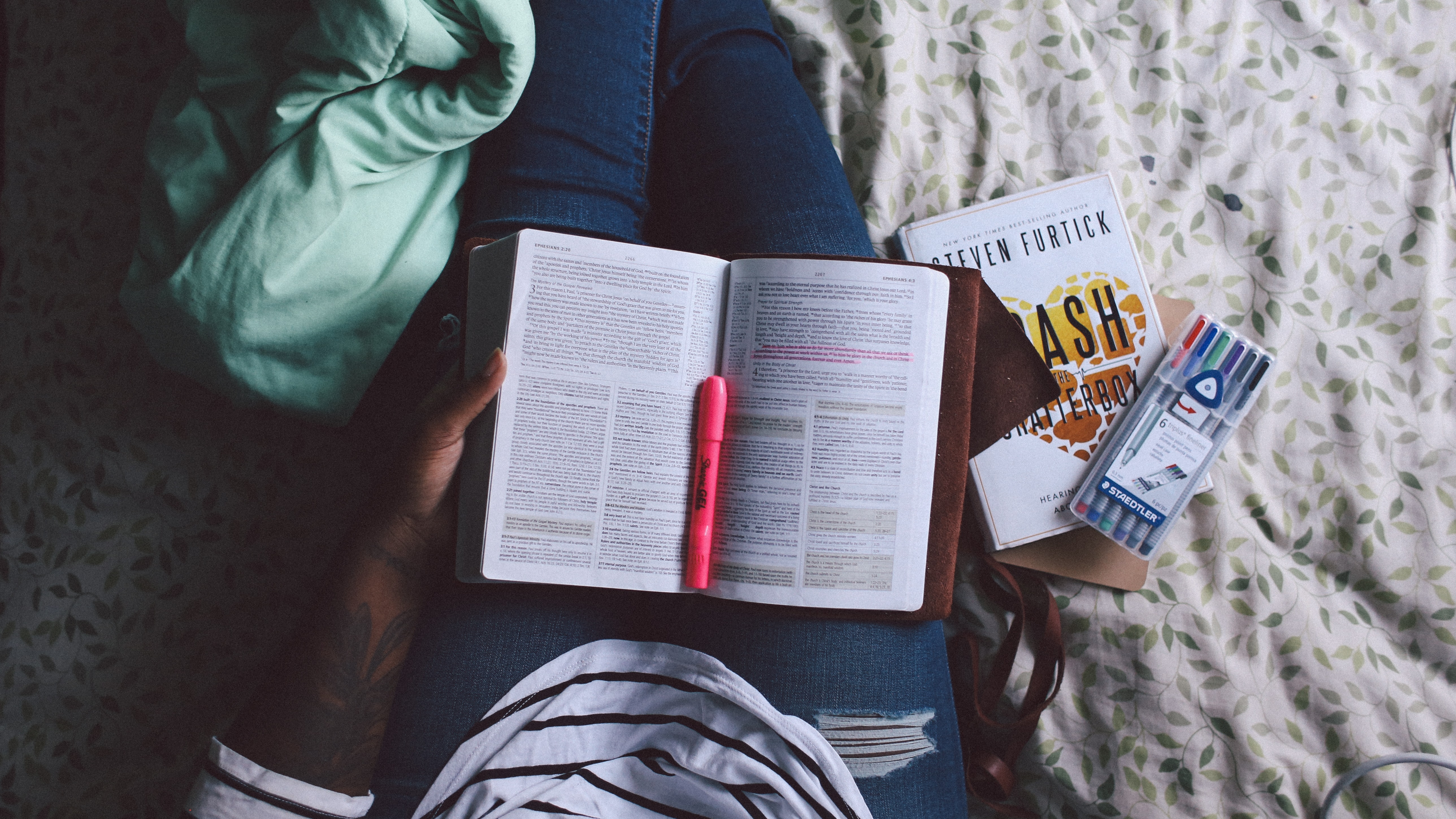 A book lies open in a person's lap, a highlighter on top of it and another book and pens at the side.