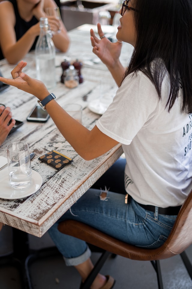 Une femme asiatique rit en gesticulant devant deux autres femmes de l'autre côté d'une table dans un café.