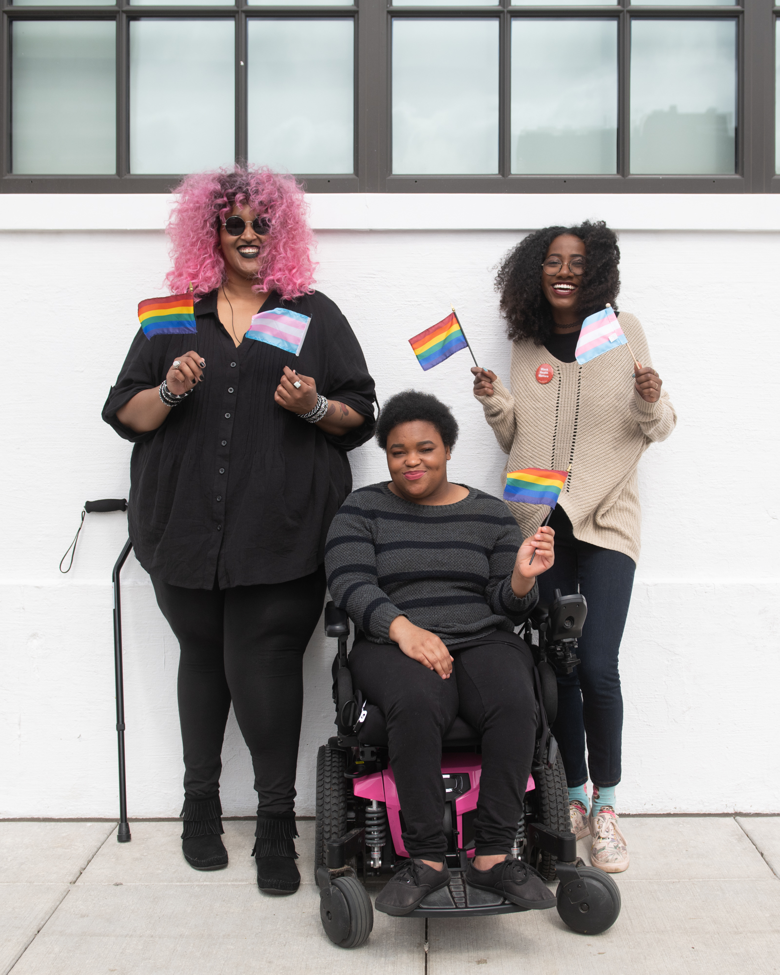 Trois personnes noires et handicapées, dont deux non binaires, sourient et brandissent des mini-drapeaux de la fierté LGBT et transgenre.