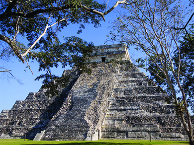 Uma fotografia mostra El Castillo, uma pirâmide escalonada com um conjunto de largos degraus de pedra subindo pela frente e uma estrutura quadrada com uma entrada no topo.