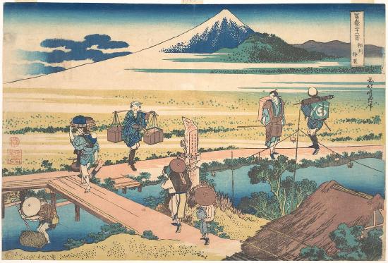 Seven people carrying goods walking on a boardwalk over a river with a mountain in the background