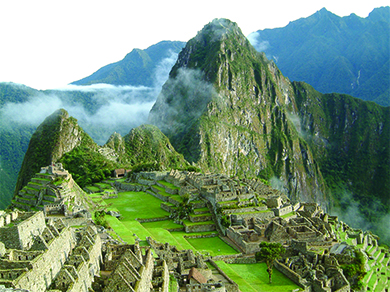 تُظهر صورة لماتشو بيتشو أنقاض مجمع من المباني ذات الجدران الحجرية والتراسات المتدرجة باللون الأخضر مع العشب والهرم، مع الجبال العالية في الخلفية.