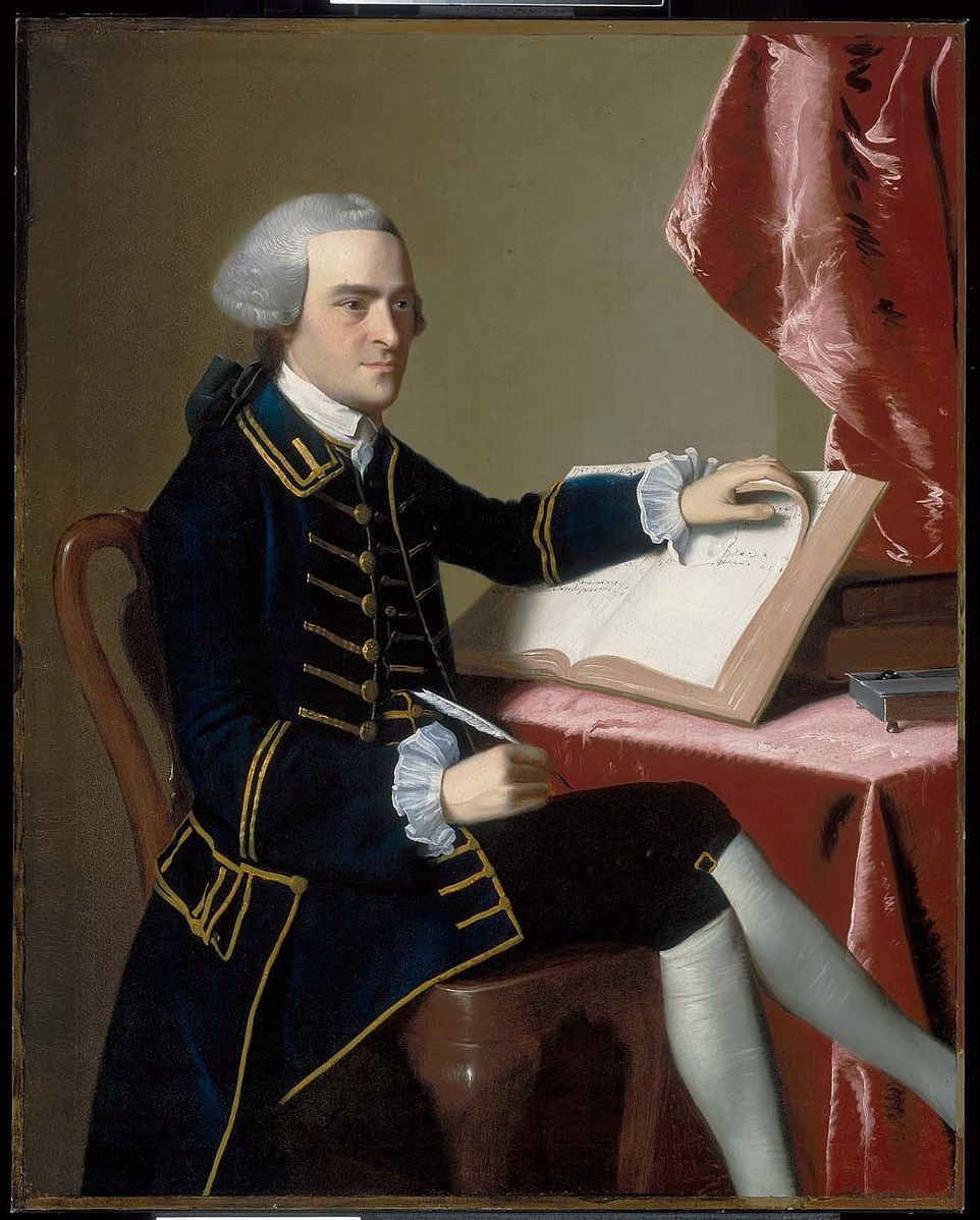 A man sitting at a desk writing in a book wearing a blue coat