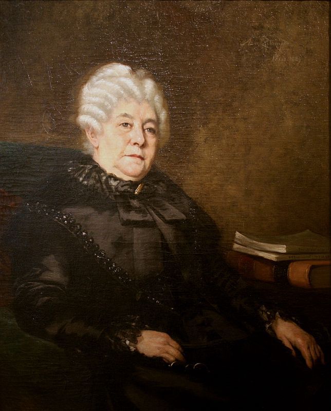 A woman wearing a black dress with grey hair sitting in a chair 