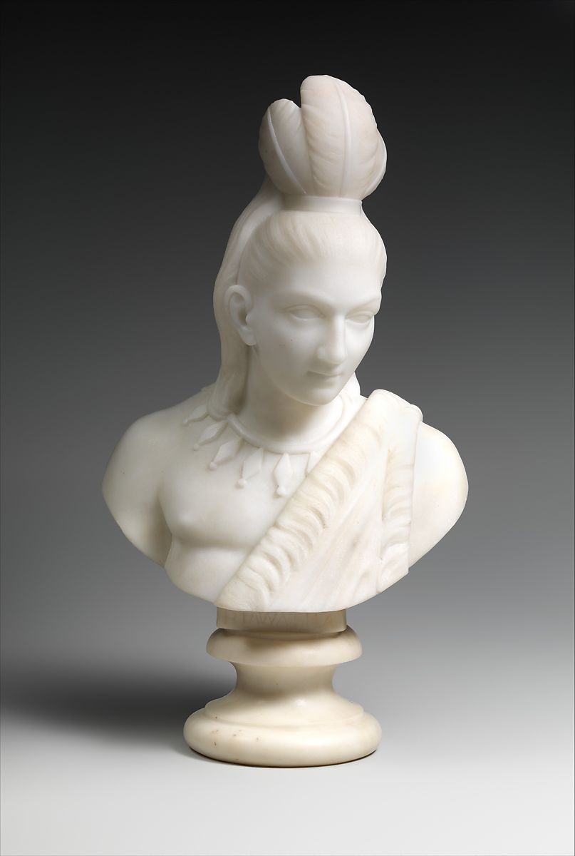 A carved marble bust of a young man with long hair and 3 feathers on the top of his head