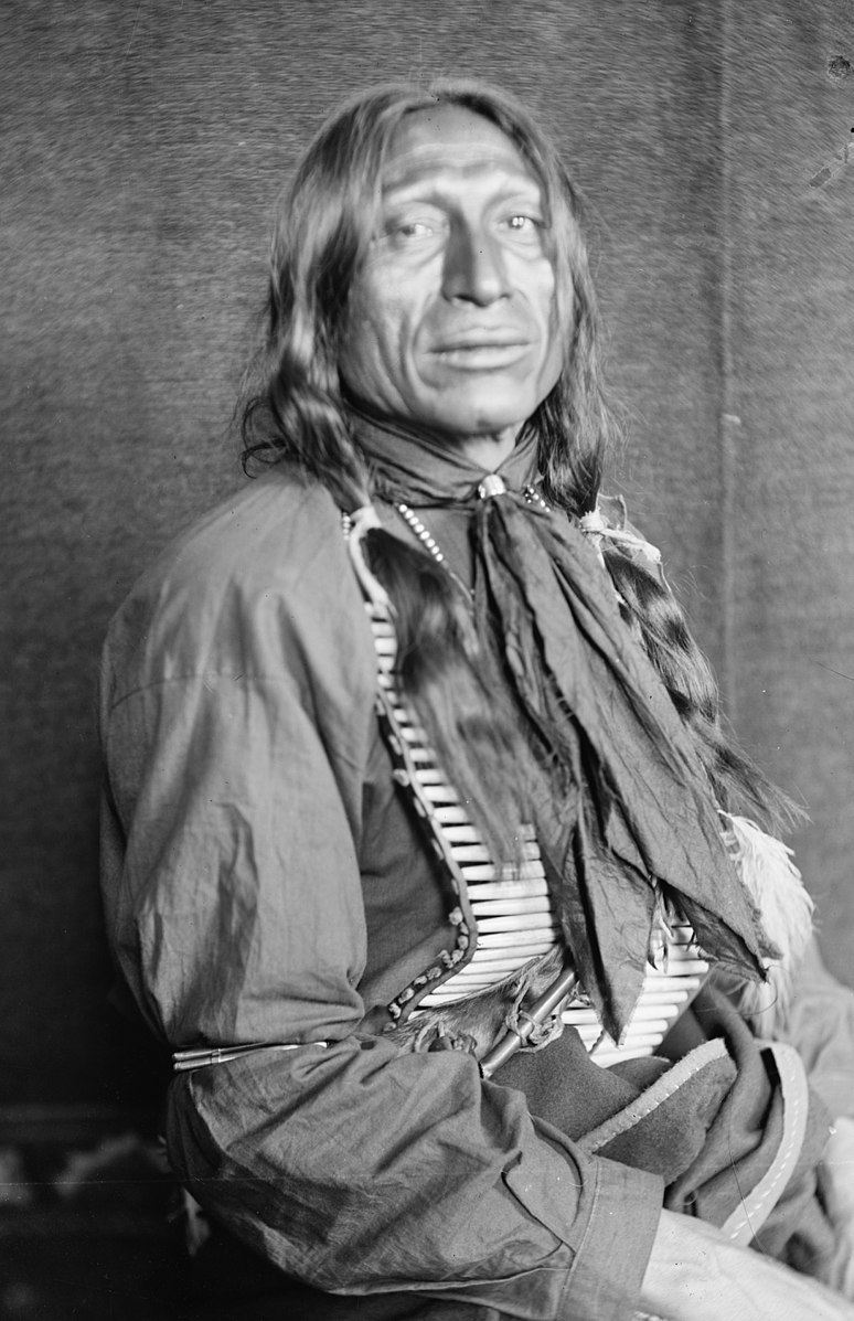 A man sitting in a chair with long hair and traditional native clothing