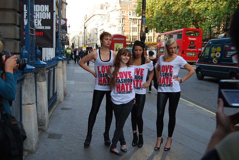 4 شباب يقفون ويبتسمون تحت لافتة تقول «أسبوع الموضة في لندن» على رصيف المدينة. جميعهم يرتدون قمصانًا مكتوب عليها «أحب الموضة وأكره المصانع المستغلة للعمال»