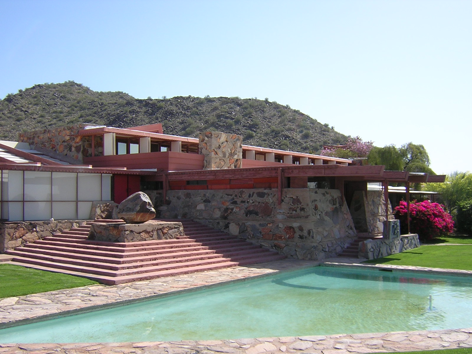 A stone house with decks and red beams with a pool at the bottom of the steps