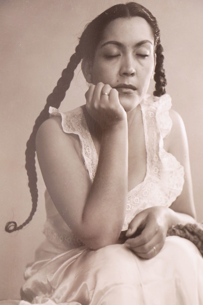 Woman sitting with her one hand in her lap and the other hand under her chin with long black braids