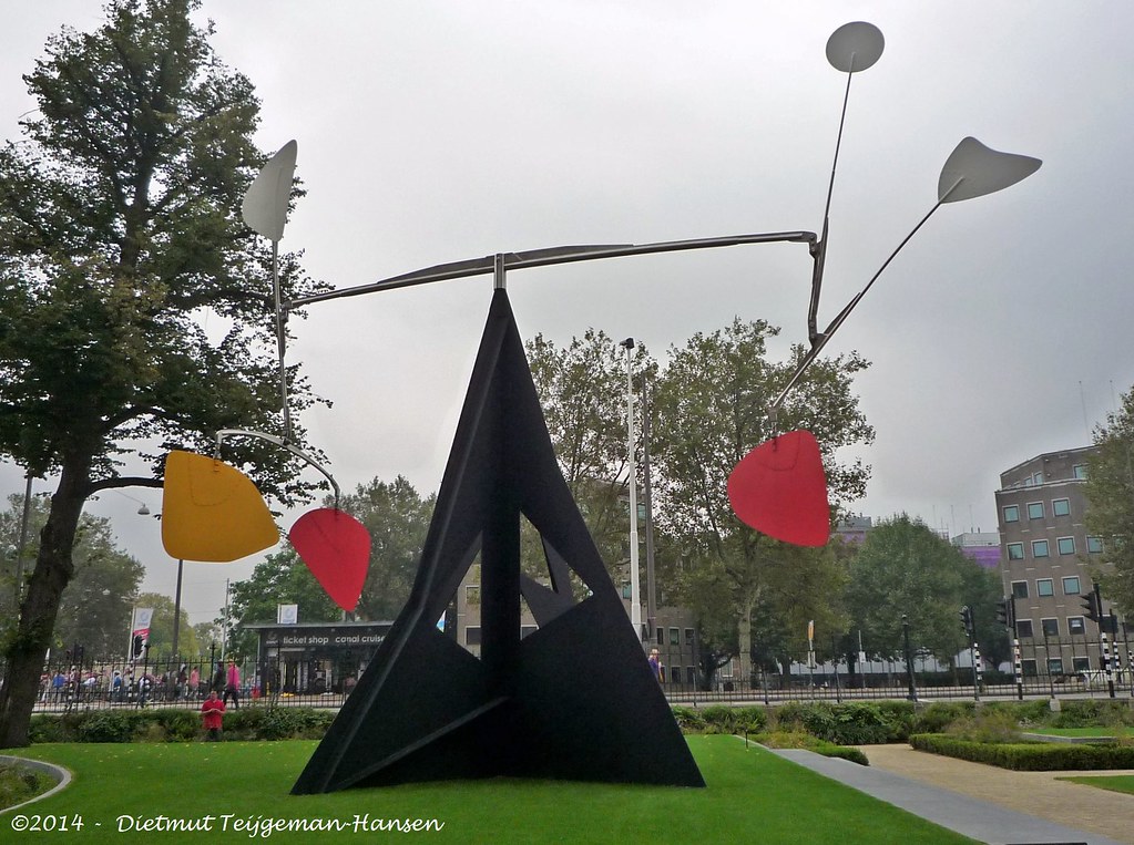 A large metal sculpture with a triangle for support in primary colors in a park