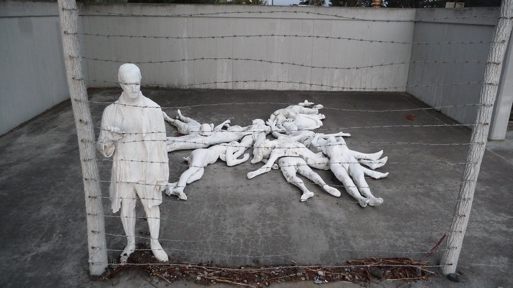 several white figures laying on the concrete while one is standing up holding on to the barb wire