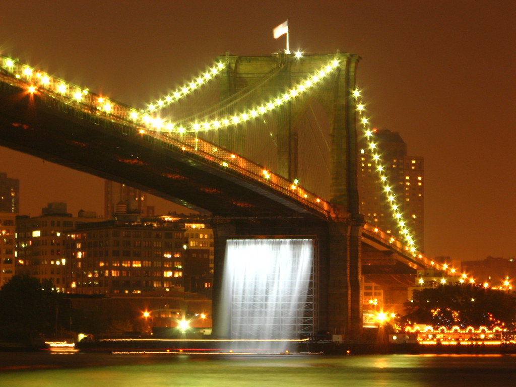 Brooklyn bridge with a water fall and lights on one of the up right supports