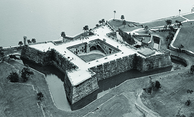 Uma fotografia aérea mostra o forte espanhol de Castillo de San Marcos, uma estrutura quadrada de paredes altas voltada para a água e incluindo um fosso ao redor.