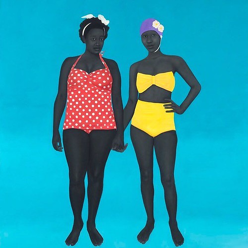 Two women standing against a blue background wearing a red bathing suit and the other is wearing a yellow bathing suit
