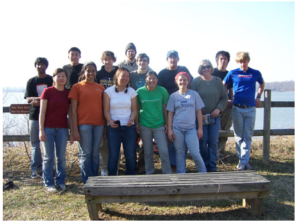 A group of students stand along a fence in front of a lake with a low bench in front of them.