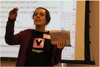 A woman wearing a black shirt decorated with a pink graphic V and "Vlogger" holds a notebook while speaking in front of a presentation screen.