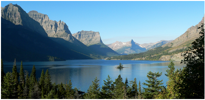 Landscape scene from Glacier National Perk depicting a lake with a small island surrounded by tall granite mountains with trees in the foreground.