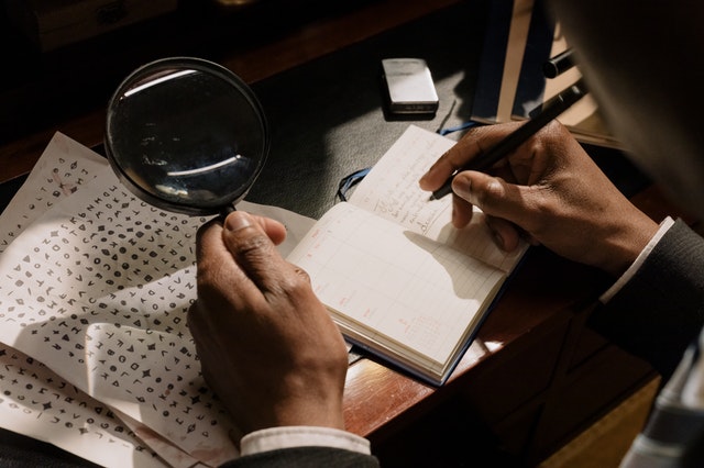 La mano marrón de un hombre sostiene una lupa sobre una página de personajes en un guión desconocido. El otro escribe notas en un libro.