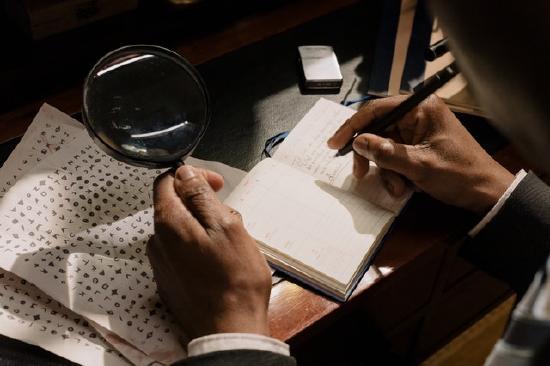 A man's brown hand holds a magnifying glass over a page of characters in an unknown script. The other writes notes in a book.