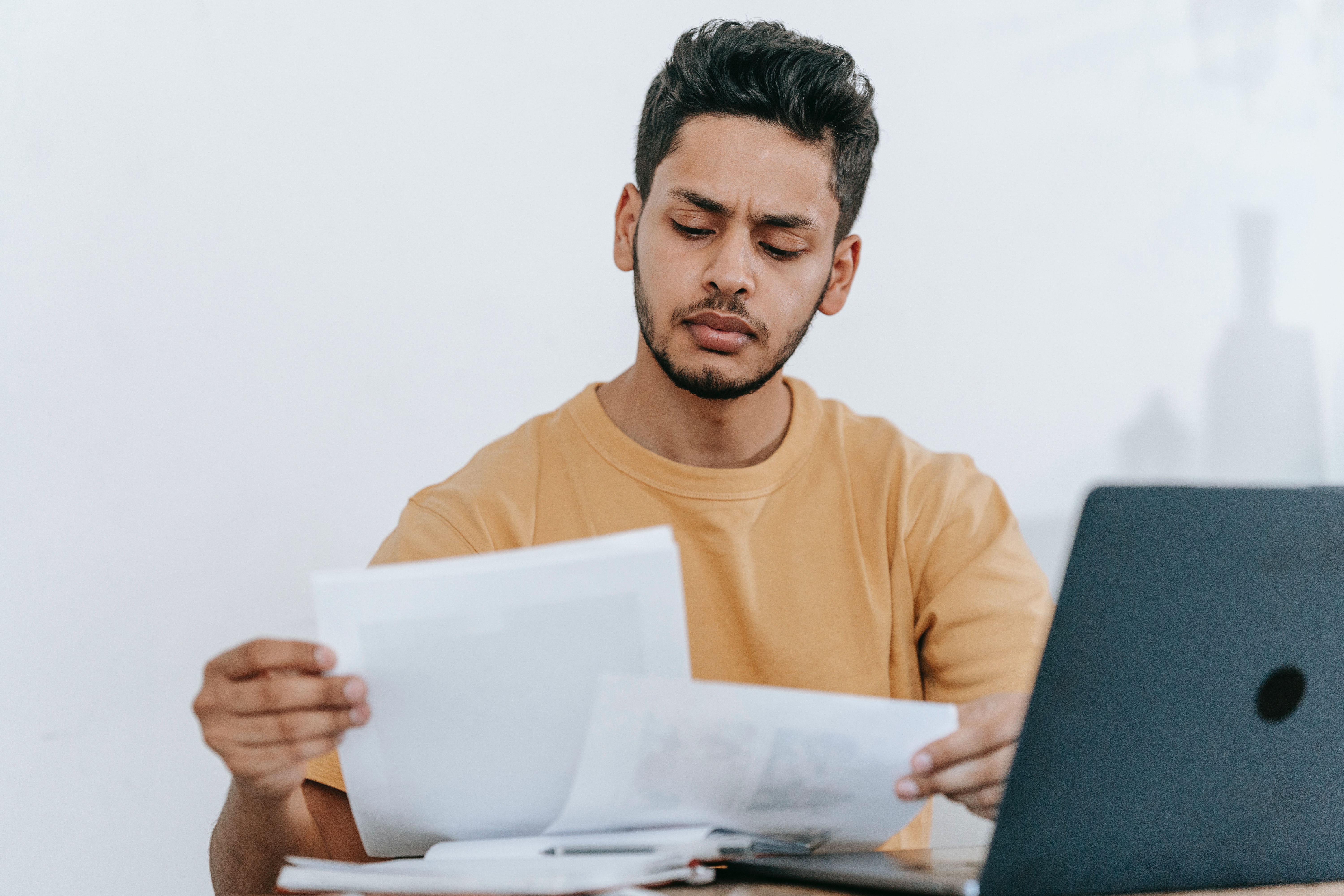 A man seated in front of an open laptop frowns thoughtfully, contemplating two pieces of paper.