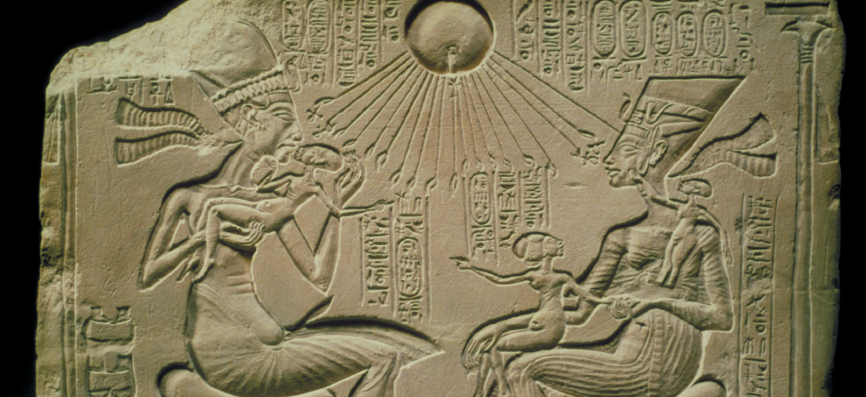 Fragment de pierre sculpté avec des images de deux adultes tenant des enfants. Au-dessus d'eux se trouve le soleil, avec des rayons qui descendent vers le bas pour toucher les adultes et les enfants. Des hiéroglyphes égyptiens entourent les images.