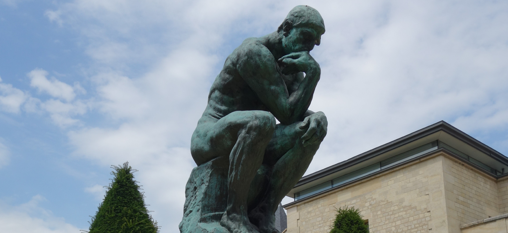 Grande escultura em bronze de um homem sentado em uma posição curvada, cotovelo no joelho e queixo na mão. Ele olha pensativamente para um espaço próximo.
