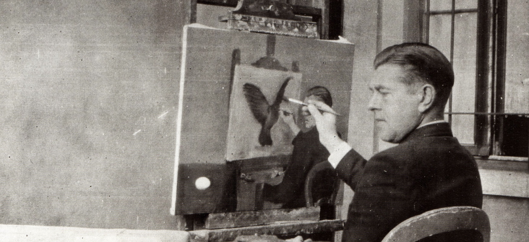 Photographie en noir et blanc d'un homme assis tenant un pinceau sur une toile presque terminée posée sur un chevalet.