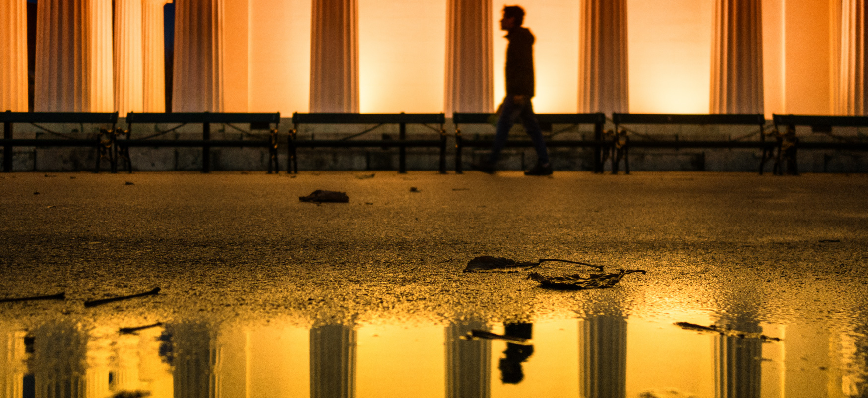 Uma pessoa andando em frente a um grande prédio forrado com colunas de mármore. O reflexo da pessoa é visível em uma poça em primeiro plano.