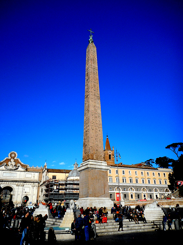 Obelisco egípcio muito alto no meio de uma praça pública. Atrás dela estão vários edifícios de pedra de quatro andares. As pessoas se sentam na base do obelisco.