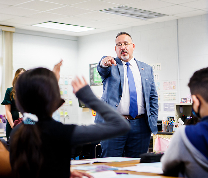 Uma sala de aula primária. Um homem vestindo terno e gravata está no centro da sala. Os alunos em primeiro plano levantam as mãos no ar, esperando que sejam chamados.