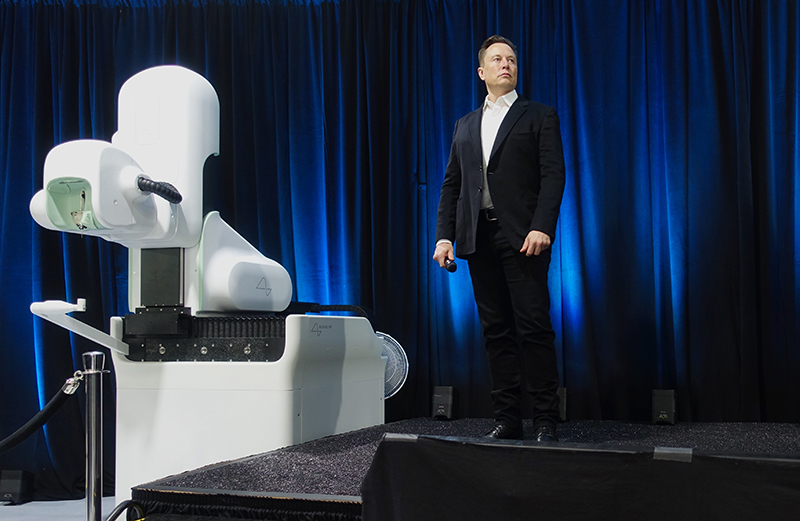 Ілон Маск стоїть на сцені біля великої, гладкої машини, з гладкими плоскими поверхнями і закругленими кутами.