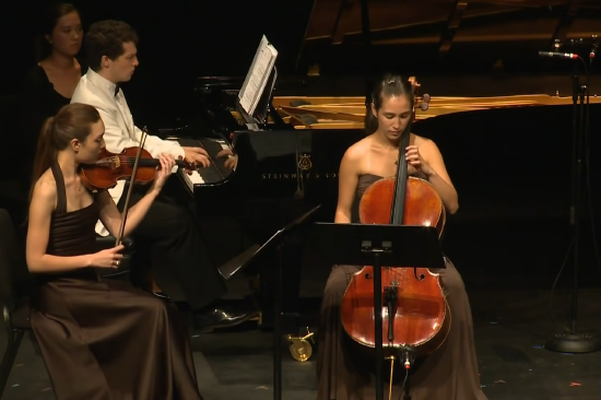 Performance of Ravel's Piano Trio featuring a violin, cello, and piano. A lonely page turner lurks in the back behind the pianist.