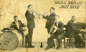 Yellowed promotional postcard featuring the original Dixieland Jass Band. The postcard shows five men each playing various instruments including: trombone, clarinet, trumpet, piano, and drums.