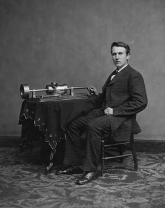 Black and white image of Thomas Edison seated next to the phonograph.