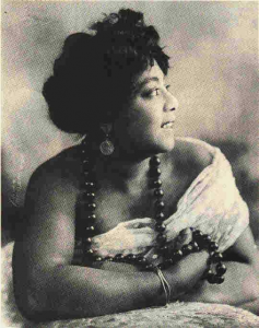 photographic image of Mamie Smith looking away from the camera, draped in a white fabric, wearing big black beads.