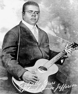 Black and white photographic image of Blind Lemon Jefferson wearing glasses, holding a guitar.