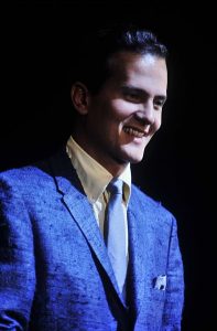 Color photographic image of Pat Boone wearing a blue jacket and blue tie with a dark background.