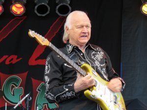 Color photographic image of Dick Dale wearing a black jacket with white lettering and holding a yellow electric guitar.