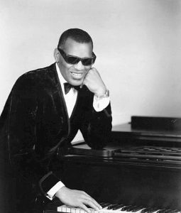 Black and white photographic image of Ray Charles sitting in a suit next to a piano wearing sunglasses.