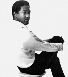 Black and white photographic image of Sam Cooke in a white shirt and black pants. He is seated away from the camera with his head turned looking back at the camera.
