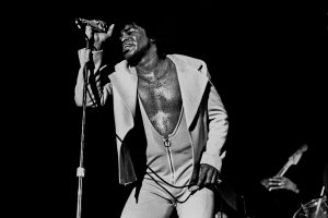 Black and white photographic image of James Brown in an open shirt with his chest exposed singing into the microphone.
