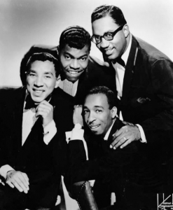 Black and white photographic image of Smokey Robinson and Miracles. There a four men in suits leaning in together looking at the camera.