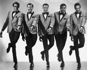 Black and white photographic image of the Temptations. There a five men in matching suits dancing together in synchronization. They have their left feet raised and the left hands pointed out towards the camera.