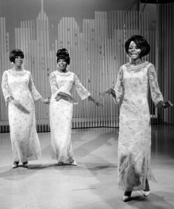 Black and white photographic image of the Supremes. There are three women in matching dresses dancing together in synchronization. The background looks like a graphic image of city scape.