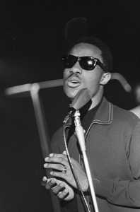 Black and white photographic image of the Stevie Wonder wearing sunglasses and singing into a microphone.