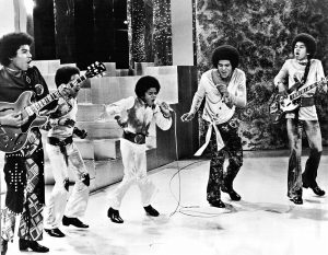 Black and white photographic image of the Jackson 5 singing in action. All of the members have afros. Two hold electric guitars. Several hold microphones.